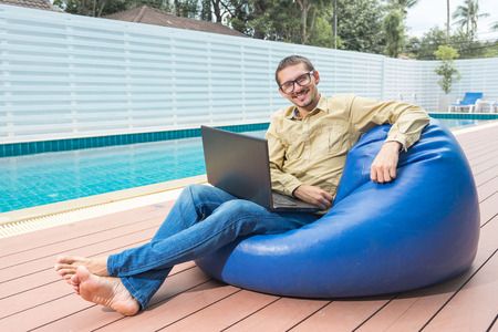Freelance businessman. Young handsome man in glasses working on laptop while sitting on the beanbag nearly swimming pool.の写真素材