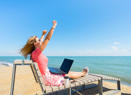 I'm the winner - young woman sitting with laptop, coffee cup and smartphone on the beach.の写真素材