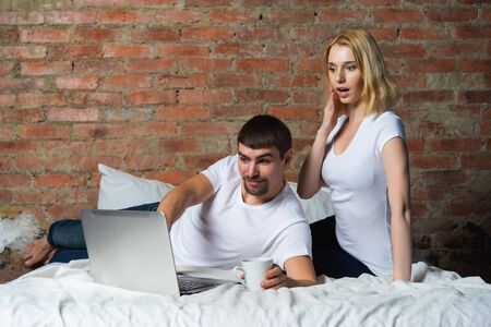 Young couple of guy and girl in bed and watching shocked news on laptop.の写真素材