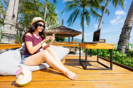 Woman sitting in beach cafe with laptop and coconut and using smartphone.の写真素材