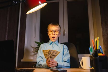 Surprised child in glasses sitting at the table with money in hands. Focus on the boy.の写真素材