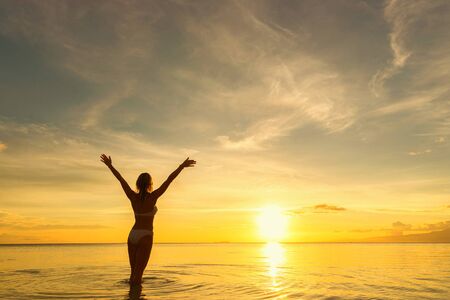Back of young woman standing in water with raised arms and looking at sea and sunset.の写真素材