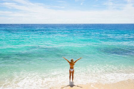 Happy young woman in white swimwear with raised hands on the beach. Back view.の写真素材