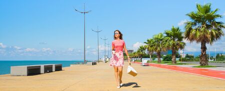 Young beautiful woman in red dress walking on the beach boulevard with coffee cup and bag. Banner edition.の写真素材