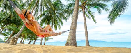 Young happy woman in hat and sunglasses laying in hammock on the tropical beach. Banner edition.の写真素材
