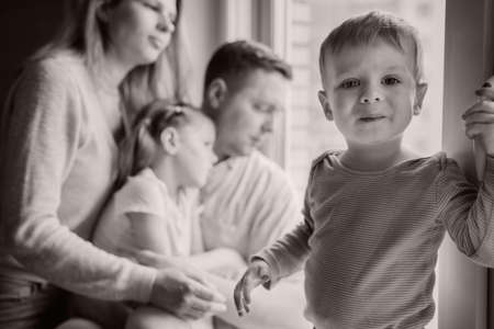 cute caucasian little boy standing on window sill. His parents and sister on background looking out the window. Home isolation during world pandemic. Black and white Image with selectie focusの写真素材