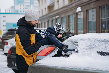 cute caucasian boy wearing scandinavian knit hat helping to brush snow from a car on winter day near...