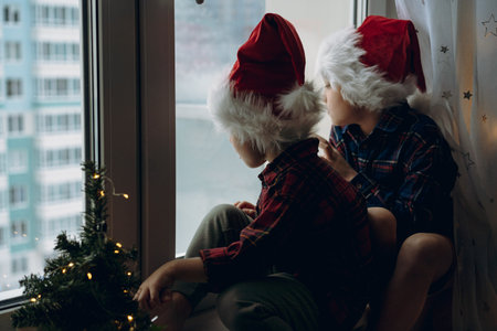 back view of cute caucasian children wearing santa hats and plaid shirts sitting hugging by the...