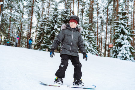 little boy learning to ride on snowboard on ski slope. Winter leisure. Pine forest backgroundの写真素材