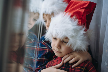 cute caucasian children wearing snta hats and plaid shirts sitting hugging by the window waiting for...