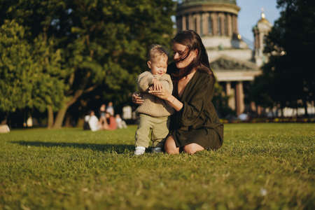 Young Caucasian lady, mother of little son sitting on grass in front of st isaac cathedral, Saint petersburg, Russia and helping boy stand holding him by hand. Image with selective focusの写真素材