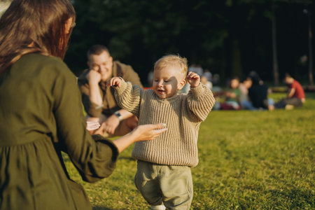 cute little caucasian boy learning to walk on lawn in park. His mother opening wide arms to catch him. Father on background, watching with tenderness. Image with selective focus. high quality photoの写真素材