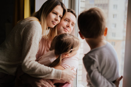 cute caucasian little boy standing on window sill. His parents and sister on background looking out the window. Home isolation during world pandemic. Image with selectie focus and toningの写真素材