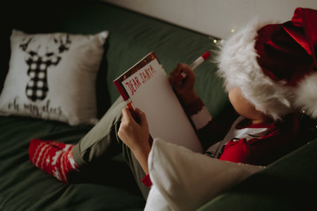 Back view of little boy in santa tee shirt and hatwriting letter to Santa Claus sitting on couch ....
