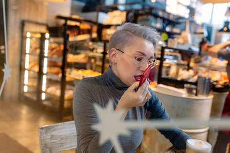 young caucasian blond woman recording audio message with smartphone sitting in cafe. View through glass, Image with selective focusの写真素材