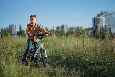 cute caucasian boy in striped henley and brown jean jacket smiling cycling on the glade of fireweed. city buildings background. Image with selective focus.の写真素材