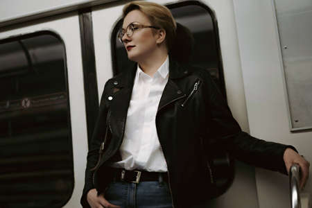 young caucasian short haired blond woman wearing leather jacket, jeans and glasses standing by door in metro carriage, looking aside. Image with selective focus. high quality photoの写真素材