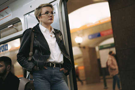 young caucasian short haired blond woman wearing leather jacket, jeans and glasses standing by door in metro carriage, looking aside. Image with selective focus. high quality photoの写真素材