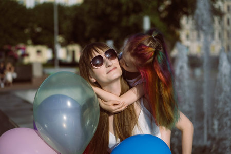 cute caucasian smiling girl with colorful dyed hair kissing mom on cheeck sitting by fountain. Image...
