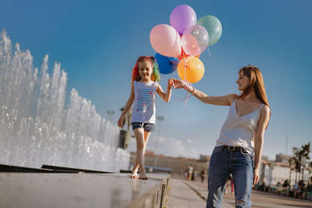 Mother walking with daughter by hand along city fountain. Little girl barefoot holding baloons. high quality photoの写真素材