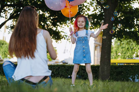 Mother and daughter having fun in park blowing bubbles. Girl holding baloons for birthday. Image with selective focusの写真素材