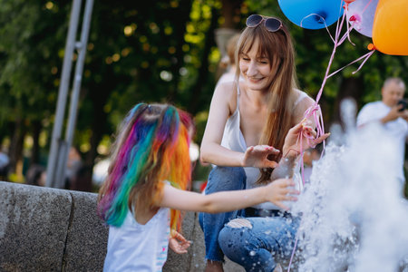 Mother and little daughter with colorful dyed hair having fun splashing near city fountain. Happy...