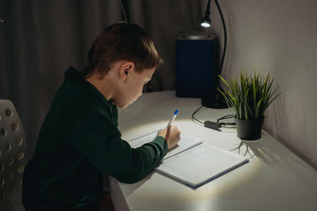 caucasian boy doing homework at home in dark room by the light of lamp. Image with selective focusの写真素材