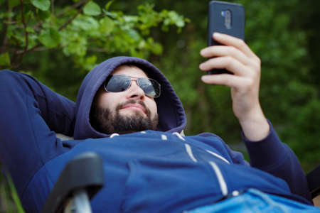caucasian bearded man wearing sunglasses having rest in camp chair with smartphone. Recreational concept. Image with selective focusの写真素材