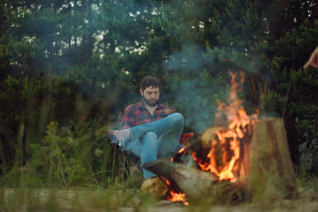 man sitting by fireplace looking with smartphone. camping concept. Image with selective focusの写真素材