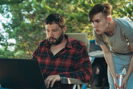 Caucasian bearded man and teenage son working wing with laptop outdoors. camping concept. parents...