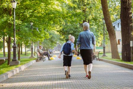 mother and son going to school holding hands. Caucasian blond boy with hairs tied up in ponytail looking at mom. Image with predictive focusの写真素材