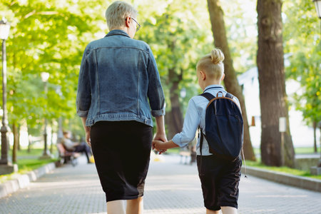 mother and son going to school holding hands. Caucasian blond boy with hairs tied up in ponytail...