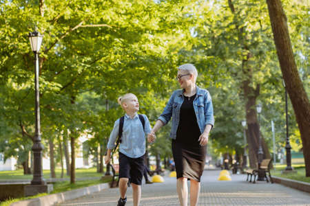 mother and son going to school holding hands. Caucasian blond boy with hairs tied up in ponytail looking at mom. Image with predictive focusの写真素材