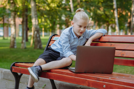 cute caucasian boy with ponytail sitting on bench in park with laptop computer. Doing homework outdoors after schoolの写真素材