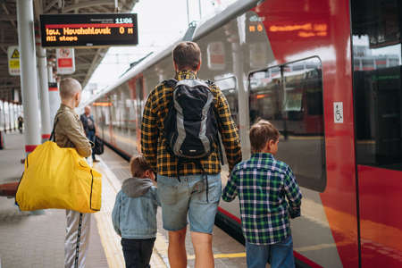 back shot of caucasian man with backpack holding children hands walking along platform going to take train.の写真素材