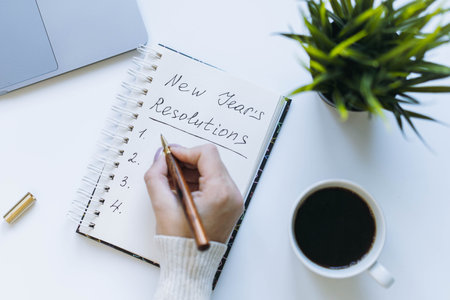 open notepad and woman hand writing new year resolutions. Flower, laptop and coffee cup on background.の写真素材