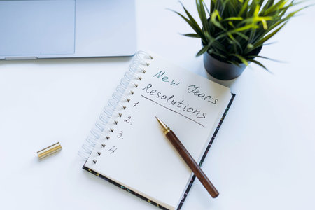 open notepad with text new year resolutions. and empty list. Flower, laptop and smartphone on...