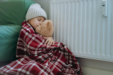 cute little boy wrapped id plaid and wearing knit hat sitting by heater hugging teddy bear. Cold at...