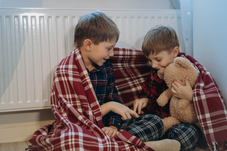 cute little boys wrapped in plaidsitting by heater hugging teddy bear, cold at home, Energy crisis. fuel povertyの写真素材