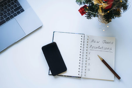 open notepad with text new year resolutions. and empty list. Christmas tree, laptop and smartphone...