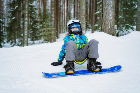 little boy sitting on snow putting his feet in snowboard bindings adjusting straps. Image with...