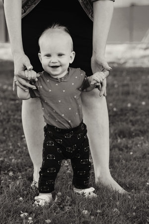 mother holding hand of baby daughter supporting child making first steps. Happy little kid learning...