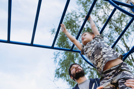 a boy doing chin-ups on monkey bars and father is helping him. Exercises on the playground on the...
