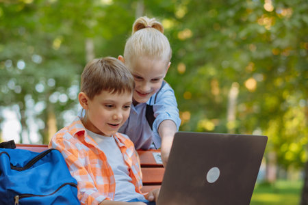 cute caucasian boys sitting on bench in park with laptop computer .Doing homework outdoors after schoolの写真素材