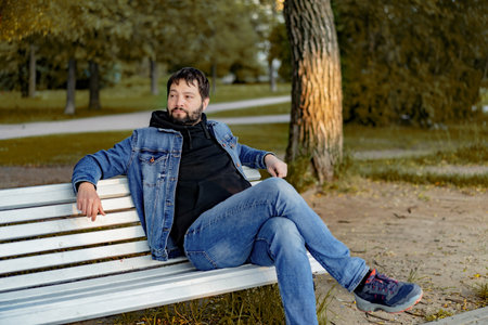 attractive caucasian bearded young man sitting on bench in park on autumn daylooking aside. high quality photoの写真素材