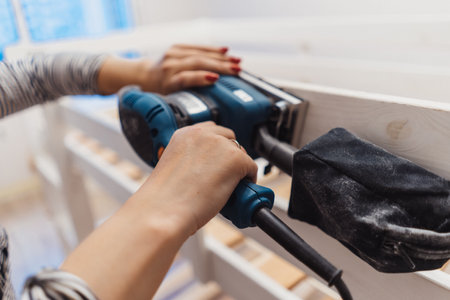 close up of woman hands rubbing wooden child bed with power sander or grinder removing old paint. Image with selective focusの写真素材