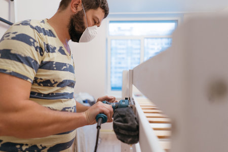 man wearing protective mask rubs wooden child bed with power sander or grinder removing old paint. Image with selective focusの写真素材
