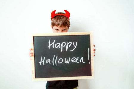 Cute little boy with red devil horns and black board with Happy Halloween text isolated on white backgroundの写真素材