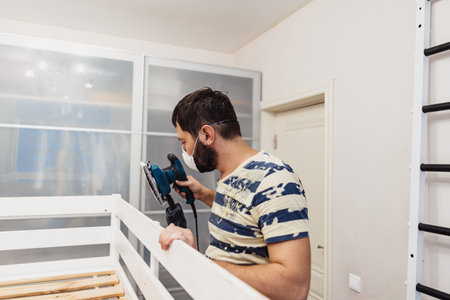 man rubs wooden child bed with power sander or grinder removing old paintの写真素材