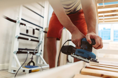 unrecognizable man wearing protective mask rubs wooden child bed with power sander or grinder removing old paint. Image with selective focusの写真素材